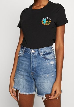 Woman wearing black t-shirt with small desert cactus embroidery and high-waisted blue denim shorts with frayed hems and distressing.