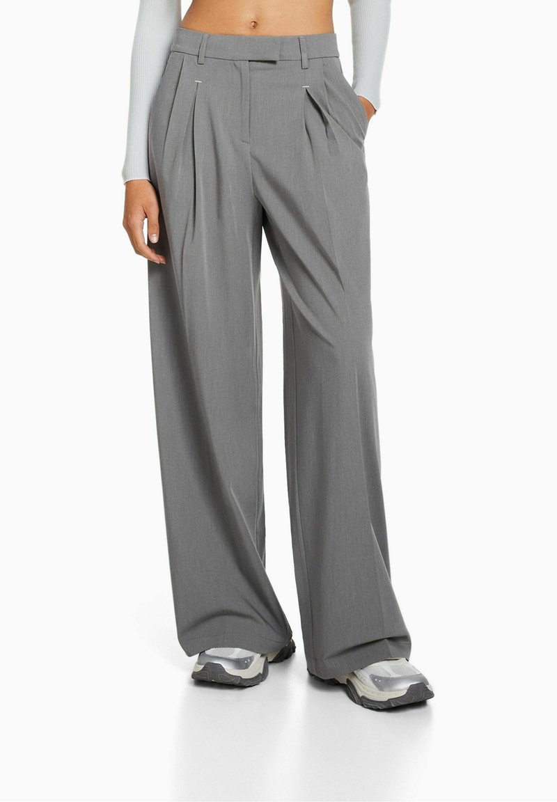 Personne portant un pantalon gris taille haute, jambes larges avec plis et des baskets, debout avec une main dans la poche sur fond blanc.