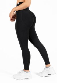 Femme portant des leggings noirs taille haute et des baskets blanches, debout avec les mains jointes, montrée de la taille aux pieds sur fond blanc.