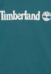 Bawełniany t-shirt w kolorze morskim z logo "Timberland" w wyraźnym, białym kolorze oraz ikoną drzewa, o gładkiej teksturze i regularnym kroju.
