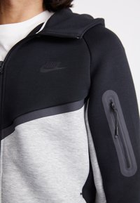 Siva in črna zadrgo zaprtih kapuc, izdelana iz mehke tkanine, z žepom na zadrgo na rokavu in diskretno Nike-ovo logo na prsih.