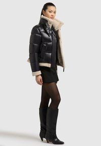 Schwarze Pufferjacke mit beigem Kunstfellfutter, Reißverschluss vorne und zwei seitlichen Taschen. Getragen mit einem schwarzen Rock und schwarzen kniehohen Stiefeln mit Absatz.