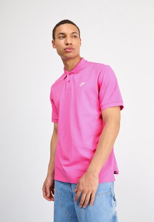 Camisa polo de Nike en un brillante color rosa, con un cuello clásico, mangas cortas y un logo blanco en el lado izquierdo del pecho. Combinada con pantalones cortos de mezclilla azul claros.