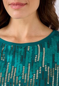 Teal top met een ronde halslijn, versierd met verticale patronen van turquoise, goud en bruine pailletten, wat een gestructureerd, glinsterend effect creëert.