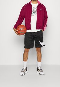 Bordeauxfärgad jacka med rosa detaljer, vit t-shirt med grafiskt tryck, svarta shorts med vit logotyp, och vita sneakers med svarta detaljer, håller en basketboll.