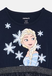 Μπλε μπλουζάκι ναυτικού χρώματος με χαρακτήρα με ξανθά μαλλιά πάνω από σχεδίαση λευκών νιφάδων χιονιού, με έμφαση σε μια λαμπερή μαύρη λεπτομέρεια στη μέση.