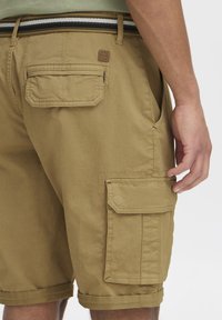 Shorts cargo beiges en tissu coton lisse. Présentent plusieurs poches, un ourlet roulé et une taille rayée avec des accents noirs et blancs.