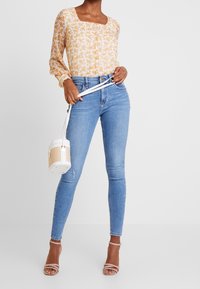 Květinová blůza s dlouhým rukávem a čtvercovým výstřihem, kombinovaná se světle modrými skinny džínami. Doplněná béžovou a bílou kabelkou přes rameno.