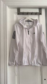 White lightweight jacket with a hood, featuring a front zipper, elastic hem, and three black stripes on each sleeve. Black logo on the chest.
