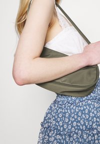 Sac à épaule vert olive avec une texture lisse et mate et une forme arrondie, porté sur un haut en dentelle blanche et une jupe à motif floral bleu.