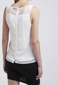 Žena v bílé bezrukávové topu se zapínáním na zádech a krajkovými detaily, v kombinaci s černou sukní, zobrazená zezadu.