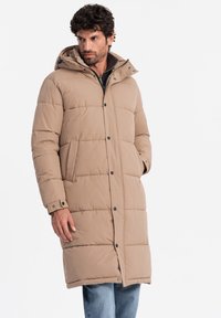 Abrigo de plumas beige con capucha, presenta un cierre frontal de botones, dos bolsillos laterales y un diseño acolchado. Lleva puesto sobre unos vaqueros azules.