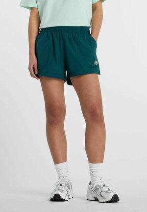 Persona che indossa pantaloncini sportivi turchesi, maglietta a maniche corte verde chiaro, calzini bianchi e sneakers New Balance bianche, in piedi con una mano in tasca.