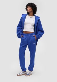 Niebieska bluza z kapturem i dopasowane dresy z miękkiego materiału; krótkie białe top; srebrne sneakersy o błyszczącym wykończeniu; szczegół z zamkiem błyskawicznym w bluzie z kapturem.