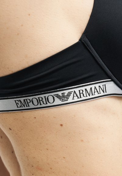 Close-up of a black Emporio Armani bra strap resting on bare skin with the brand logo and name on the elastic band.