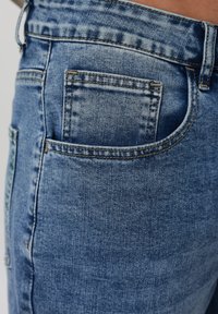 Κοντινό πλάνο μπλε τζιν παντελονιού με ορατή πίσω τσέπη, ραφές και θηλιά ζώνης στο ισχίο ενός ατόμου.