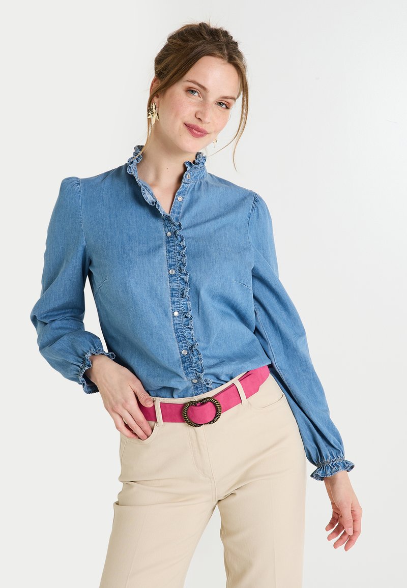 Chemise en denim bleu clair avec col et poignets froncés, boutons sur le devant, associée à un pantalon beige et une ceinture rose avec une boucle ronde.