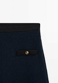 Tissu bleu marine texturé avec ceinture élastique noire et petite languette noire tricotée ornée d’un bouton rond doré.