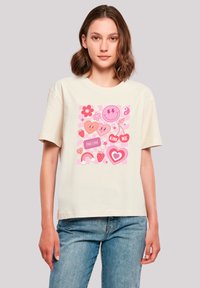 Cream-colored t-shirt featuring a pink graphic design with hearts, flowers, emojis, and a rainbow, set against a light backdrop.