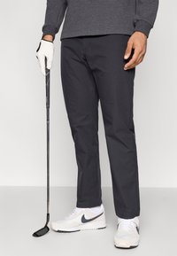 Γκρι αθλητικό παντελόνι από απαλό ύφασμα, στενή εφαρμογή, σε συνδυασμό με λευκά αθλητικά παπούτσια με μαύρο λογότυπο, κράτηση μπαστουνιού του γκολφ με γαντζωμένο χέρι.