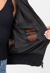 Intérieur noir de bomber avec doublure lisse, une étiquette logo orange bien visible et des poignets en molleton. Étiquettes avec instructions d'entretien visibles.