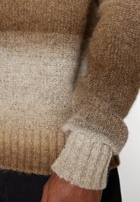 Dzianinowy sweter w gradientowych odcieniach brązu do beżu, z prążkowanymi mankietami i miękką, teksturowaną tkaniną. Zbliżenie na rękaw.