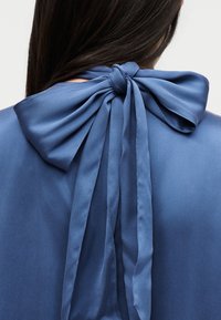 Μπλε σατέν φιόγκος δεμένος στον πίσω λαιμό ενός ενδύματος με μακριές ουρές, φορεμένος από άτομο με μακριά σκούρα μαλλιά.