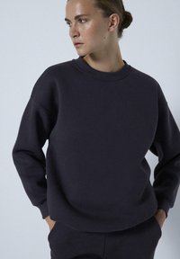 Donkergrijze sweatshirt met een ronde halslijn, zachte textuur, verlaagde schouders en geribde manchetten, gedragen met handen in de zakken.