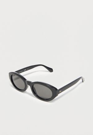 Czarne owalne okulary przeciwsłoneczne z grubymi oprawkami i ciemnymi szkłami leżące na jasnej powierzchni.