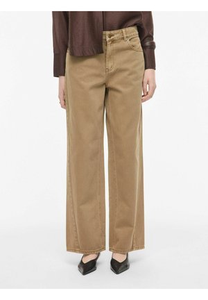 Kvinde iført beige bukser med højtaljede vide ben, brun langærmet top og sorte spidse sko, stående mod en ensfarvet baggrund.