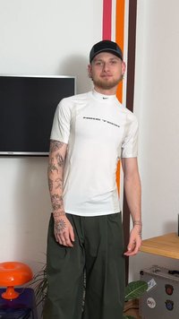 Valkoinen lyhythihainen urheilupaita, jossa on korkea kaulus, musta "NIKE TECH" -logo ja teksturoidut paneelit. Yhdistetty väljiin tummanvihreisiin housuihin.