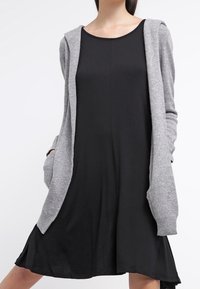 Cardigan grigio a maglia con cappuccio sopra un vestito nero a maniche corte. Il cardigan presenta tasche e un design a frontale aperto. Tessuto liscio e casual.