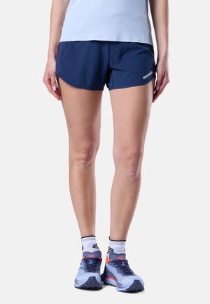 Žena v tmavě modrých sportovních šortkách značky Rossignol, bílých a černých kotníkových ponožkách a modrých běžeckých botách s oranžovými detaily, stojící na bílém pozadí.