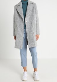 Cappotto grigio in lana con colletto a rever, due tasche frontali, bottoni doppi, indossato su un maglione teal e pantaloni azzurri chiari, abbinato a sneakers bianche.