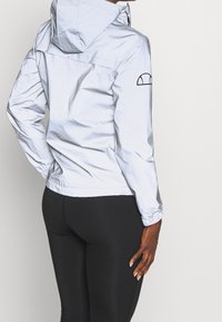 Lekka, odblaskowa kurtka z kapturem w kolorze srebrnym. Posiada elastyczne mankiety oraz czarne logo na lewym rękawie. Prosty design z minimalną ilością detali.