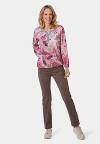 Bluse mit floralem Muster in Pink und einem blauen Ausschnitt, langen Ärmeln, kombiniert mit braunen Hosen und grauen Sneakers auf einem weißen Hintergrund.