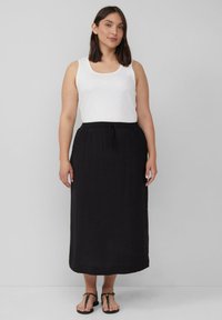 Zwarte maxi-rok met een trekkoord in de taille, gecombineerd met een witte tanktop. De stof lijkt lichtgewicht en gestructureerd. Platte sandalen maken de look compleet.