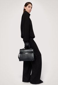 Černá kožená kabelka s krokodýlím vzorem, strukturovaného tvaru a krátkým uchy. Modelka má na sobě černý outfit a rukavice.