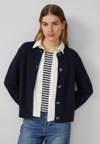 Marineblauer Strickcardigan mit Rippstruktur, runder Halslinie und Holzknöpfen. Über einem gestreiften Hemd und blauen Jeans getragen.