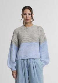 Egy nő szürke és világoskék kötött pulóvert, valamint laza világoskék nadrágot visel, és egy egyszínű világos háttér előtt áll.