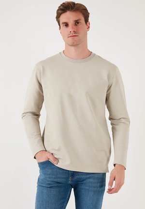 Beige long-sleeve shirt with a rounded neckline, made of smooth fabric, worn with blue jeans. Subtle stitched detailing on seams.