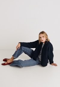 Marineblauer Strickcardigan mit abgerundetem Saum, zwei Taschen, kombiniert mit einem gestreiften Shirt und hellblauen High-Waist-Jeans sowie roten Ballerinas.