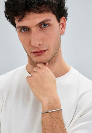 Bracciale d'argento con una fila di pietre trasparenti, indossato su un polso che riposa sotto un mento. Il modello ha i capelli corto e riccio e indossa una maglietta bianca.