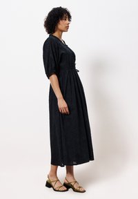 Schwarzes Maxikleid mit strukturiertem Blumendesign, kurzen Ärmeln, Kordelzug in der Taille und lockerer Passform. Kombiniert mit olivgrünen Riemchensandalen.