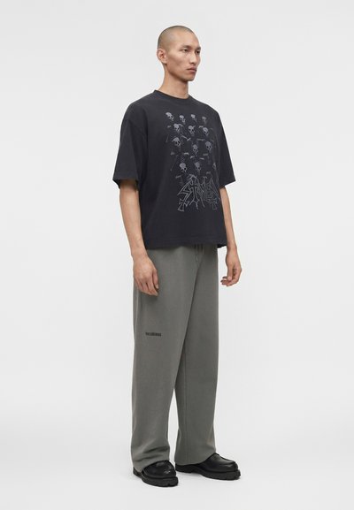 Mann steht leicht nach rechts gewandt, trägt ein übergroßes schwarzes T-Shirt mit Totenköpfen und graue Hosen mit weitem Bein sowie schwarze Schuhe vor weißem Hintergrund.