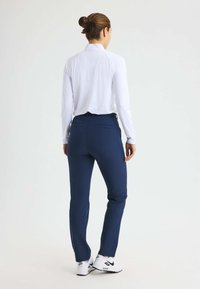 Kvinna med hår i knut, iklädd en vit långärmad topp, marinblå byxor och vita Nike-sneakers, står vänd bort från kameran mot en vit bakgrund.