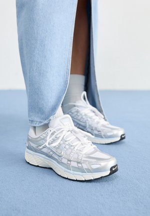 Osoba nosząca srebrno-białe sneakersy Nike z białymi skarpetkami i jasnoniebieskimi dżinsami stoi na niebieskiej podłodze.