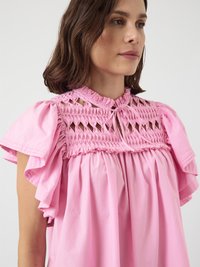 Femme avec des cheveux bruns mi-longs portant un blouse rose avec des manches courtes à volants et un encolure smockée à découpes nouée au cou.