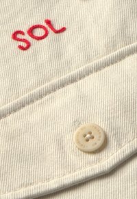 Beige stof met een gestructureerd oppervlak, met een rode geborduurde "SOL" en een crèmekleurige knop met de inscriptie "THINKING MU".