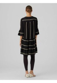 Robe noire avec un design en couches et des manches volantes. Présente des broderies en rayures argentées sur le tissu. Portée avec des leggings sombres et des chaussures.
